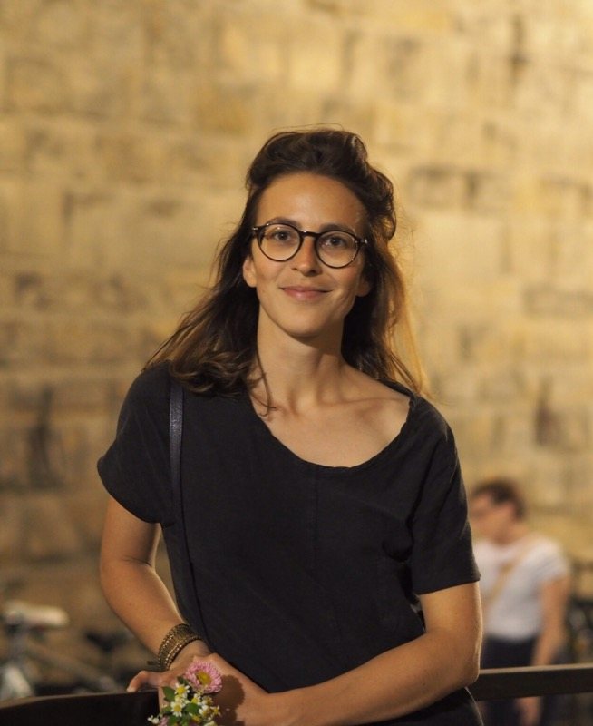 Portrait de Constance Bet, souriante, lunettes rondes, devant un mur de pierre